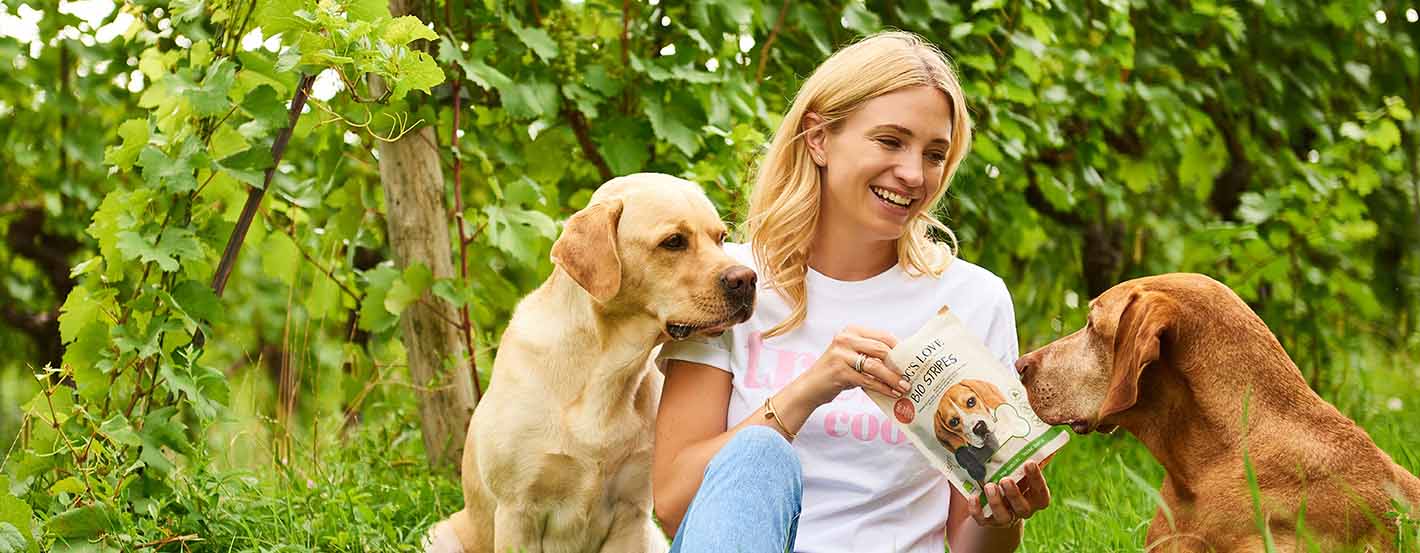 Katharina Miklauz sentada entre las viñas dando de comer a los perros Enzo y Pluto con nuestros DOG'S LOVE Snacks